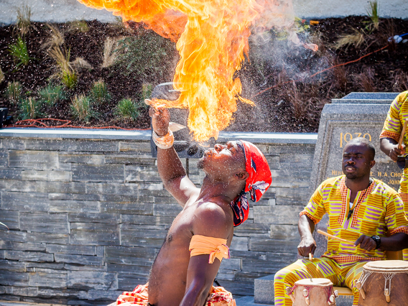 A performer at Llangollen International Eisteddfod breathing fire into the air with another performer playing a traditional African drum behind them